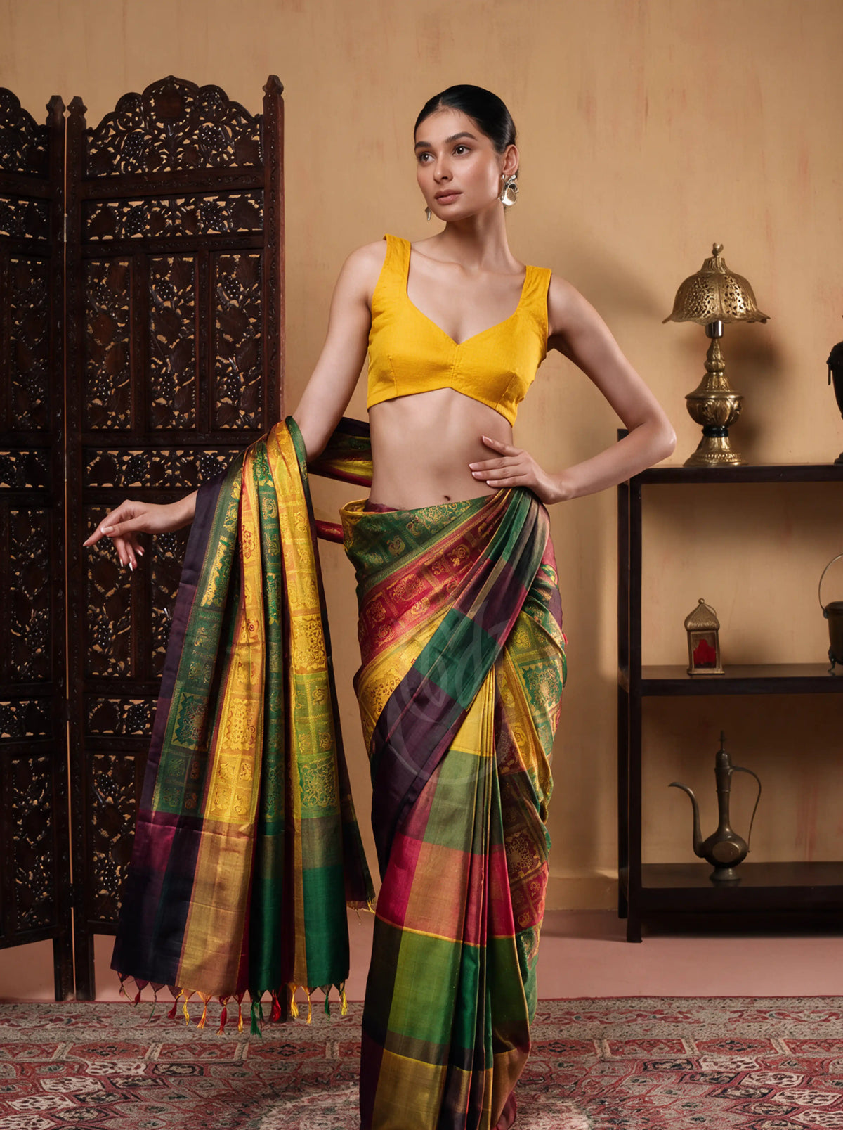 Full-length view of a model posing in a handwoven multicolor brocade silk saree with emerald green, yellow, crimson, and violet panels, paired with a bright yellow sleeveless blouse beside a wooden screen.