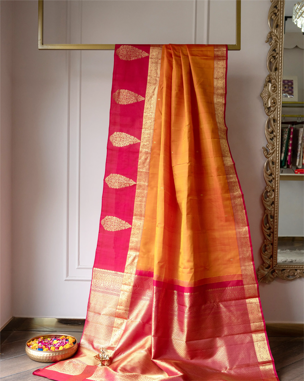 A traditional Indian silk saree draped elegantly, featuring a golden-yellow body, a bright pink border with gold zari motifs, and a wide gold zari pallu, displayed indoors against a neutral wall with a bowl of flower petals in the foreground.