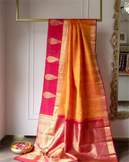 A traditional Indian silk saree draped elegantly, featuring a golden-yellow body, a bright pink border with gold zari motifs, and a wide gold zari pallu, displayed indoors against a neutral wall with a bowl of flower petals in the foreground.
