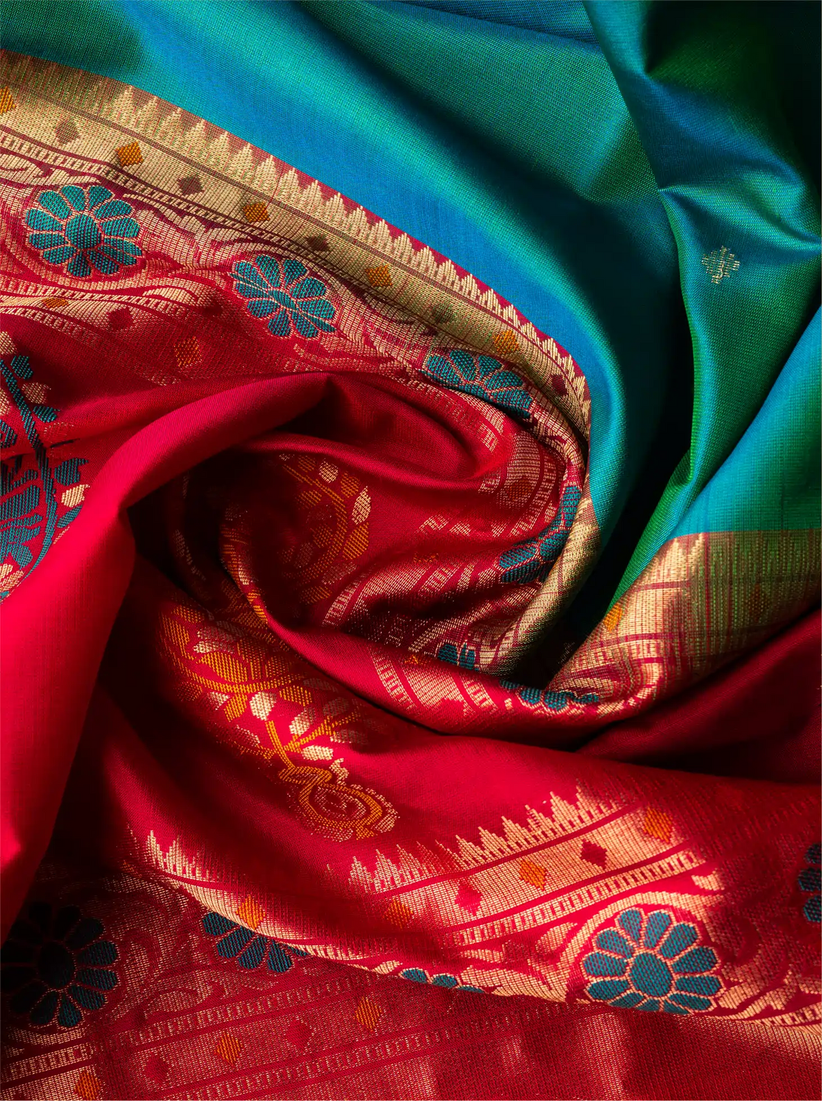 Close-up top view of a swirling handwoven Paithani silk saree showcasing a lustrous peacock teal green fabric draped alongside a vibrant red pallu intricately decorated with golden zari weaving, featuring geometric borders, floral patterns, and peacock motifs.