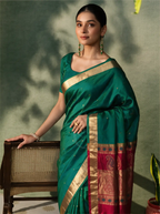 Model posing in a handwoven peacock teal green Paithani silk saree featuring small woven gold motifs on the body, paired with a broad crimson red border adorned with intricate gold zari weaving. She wears a matching scoop-neck teal green blouse with gold border sleeves, green drop earrings, and resting her hand on a wooden cane chair.