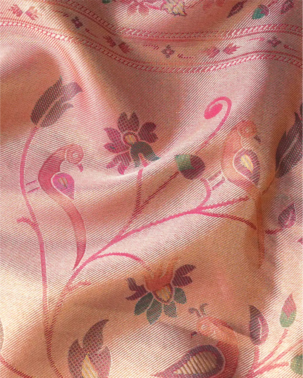Close-up shot of a peach-pink tissue silk saree showing shimmering weave with intricate pink, purple, and green zari floral and peacock motifs.
