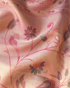 Close-up shot of a peach-pink tissue silk saree showing shimmering weave with intricate pink, purple, and green zari floral and peacock motifs.