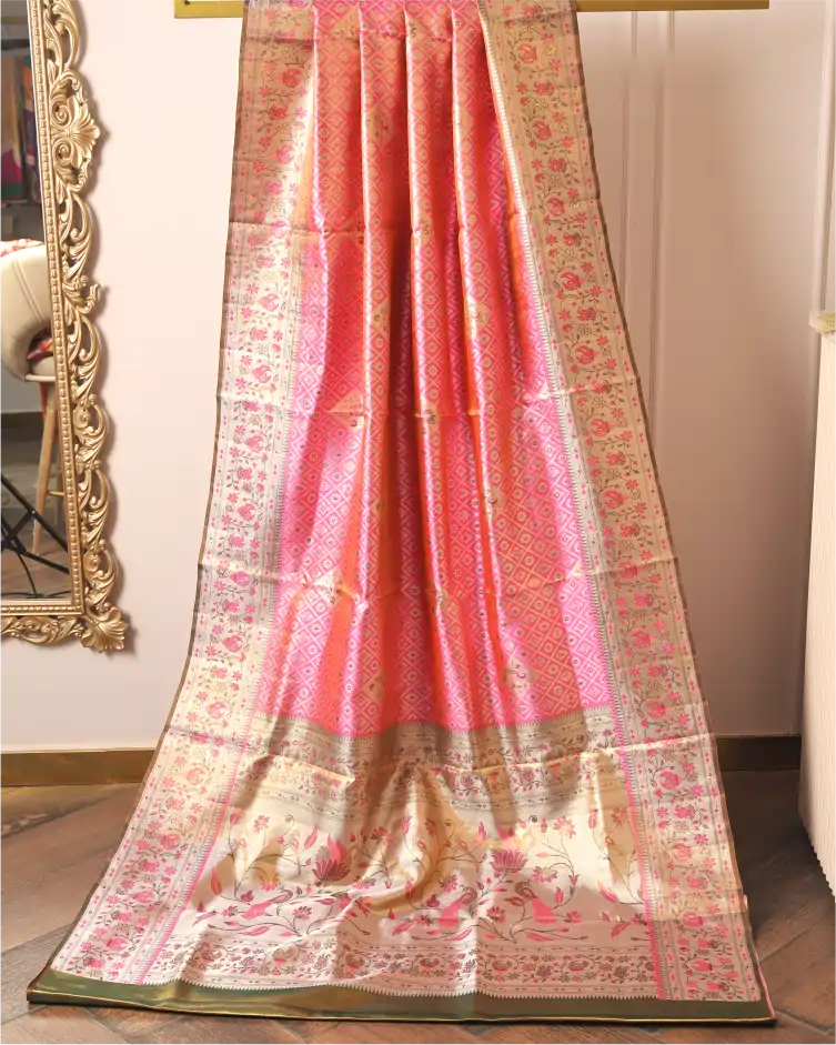 Handwoven pink tissue Patola Kanjivaram silk saree with intricate floral motifs and silver zari border draped indoors next to a mirror.