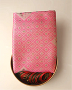 Folded pink tissue Patola Kanjivaram silk saree with woven silver zari motifs presented on a brass tray alongside green and red glass bangles.