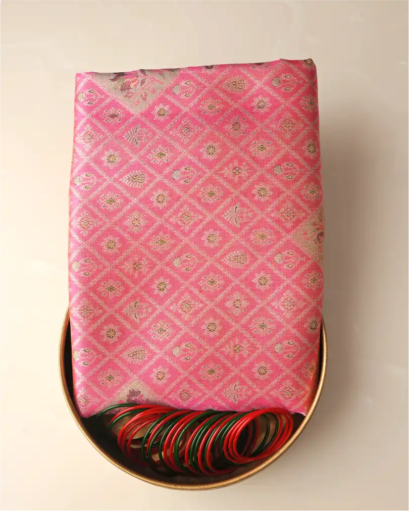 Folded pink tissue Patola Kanjivaram silk saree with woven silver zari motifs presented on a brass tray alongside green and red glass bangles.