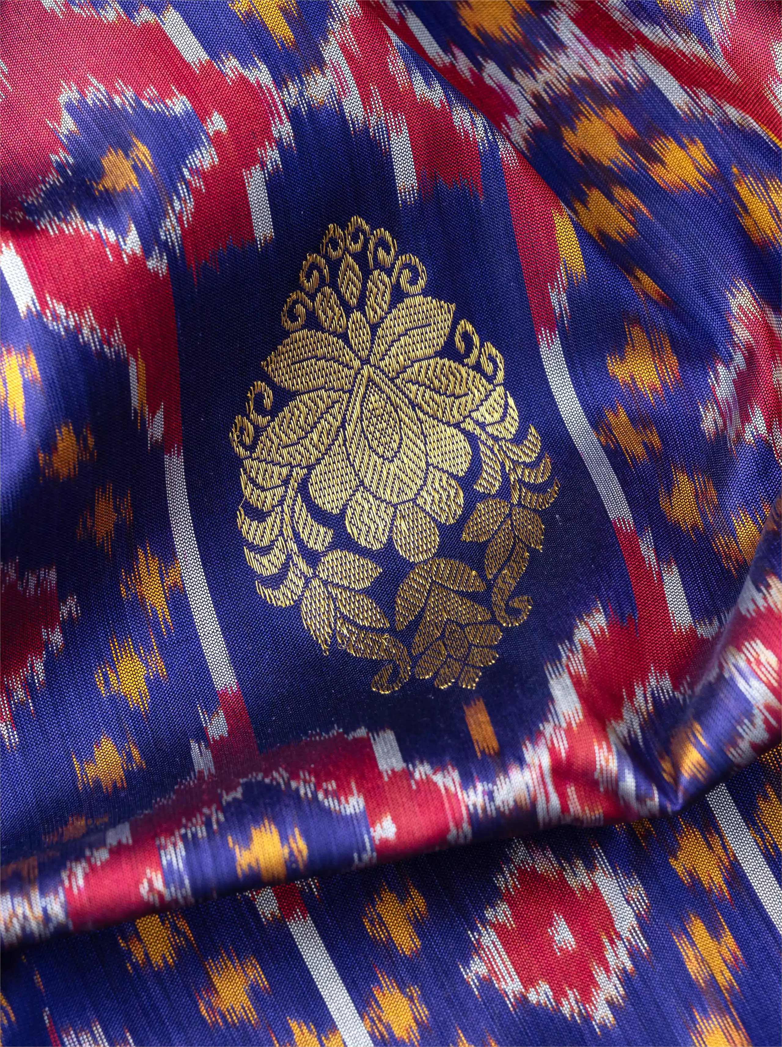 Macro shot of handwoven purple Patola silk saree showing a prominent metallic gold zari floral brocade motif over colorful ikat patterns.