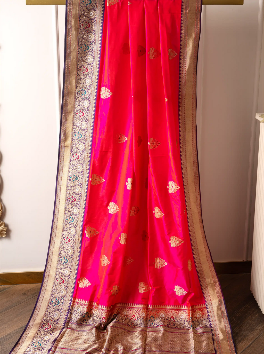 Handwoven red Katan silk saree with gold zari floral motifs and a detailed gold zari border draped on a wooden hanger.