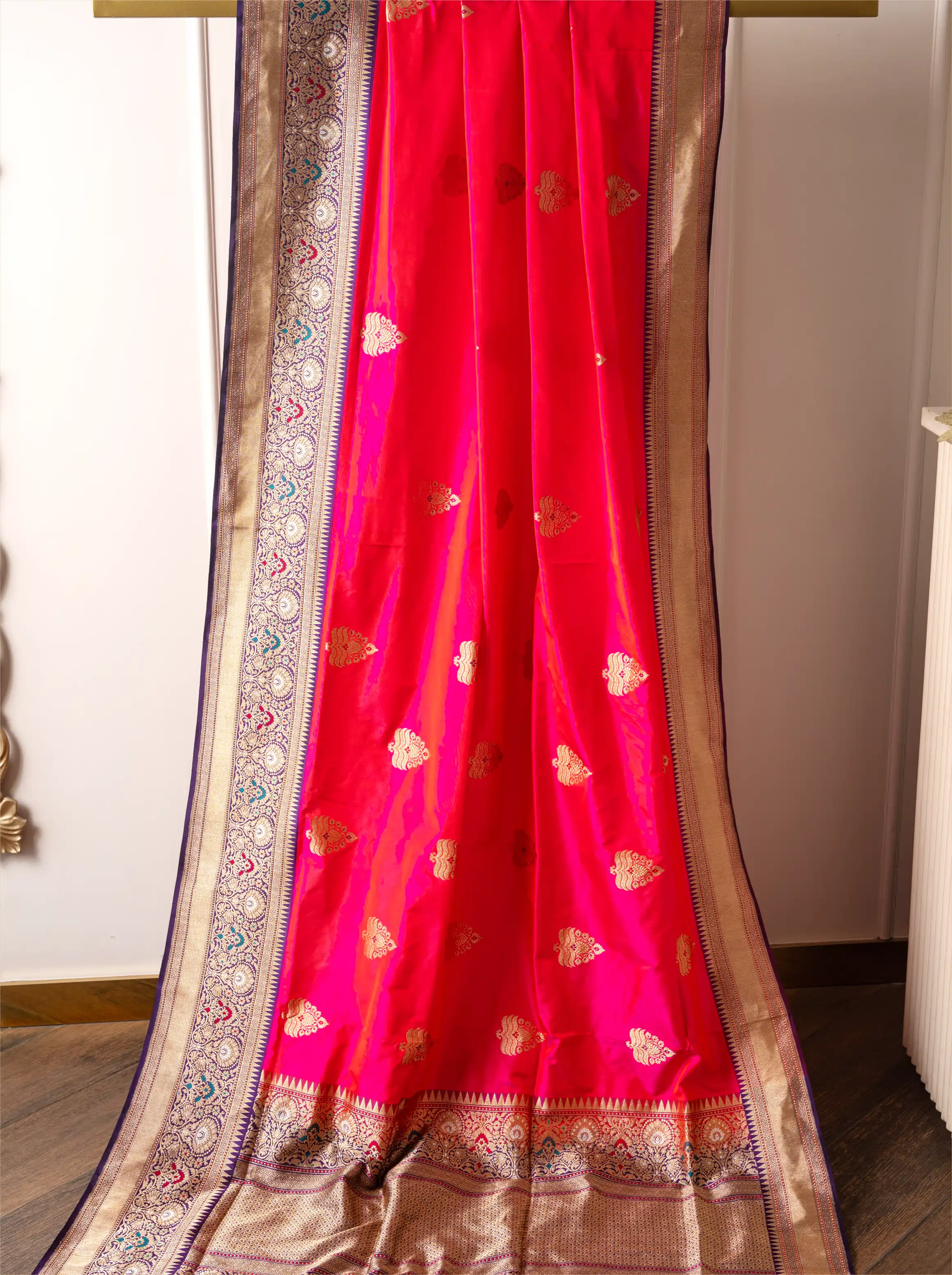 Handwoven red Katan silk saree with gold zari floral motifs and a detailed gold zari border draped on a wooden hanger.