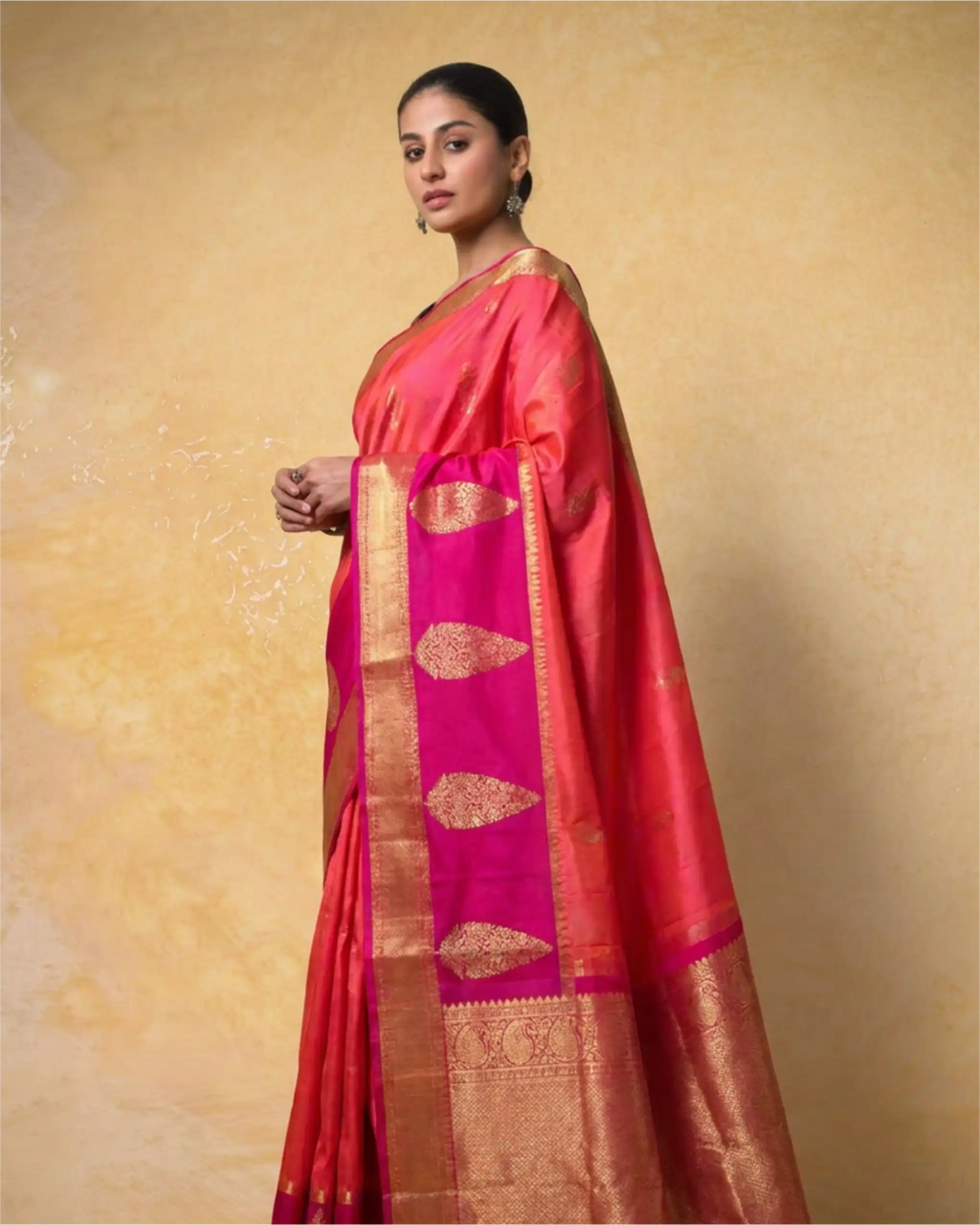 A woman modeling a vibrant pink and red pure Kanjivaram silk saree with a wide gold zari border and traditional buta motifs, set against a textured beige background