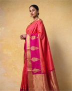 A woman modeling a vibrant pink and red pure Kanjivaram silk saree with a wide gold zari border and traditional buta motifs, set against a textured beige background