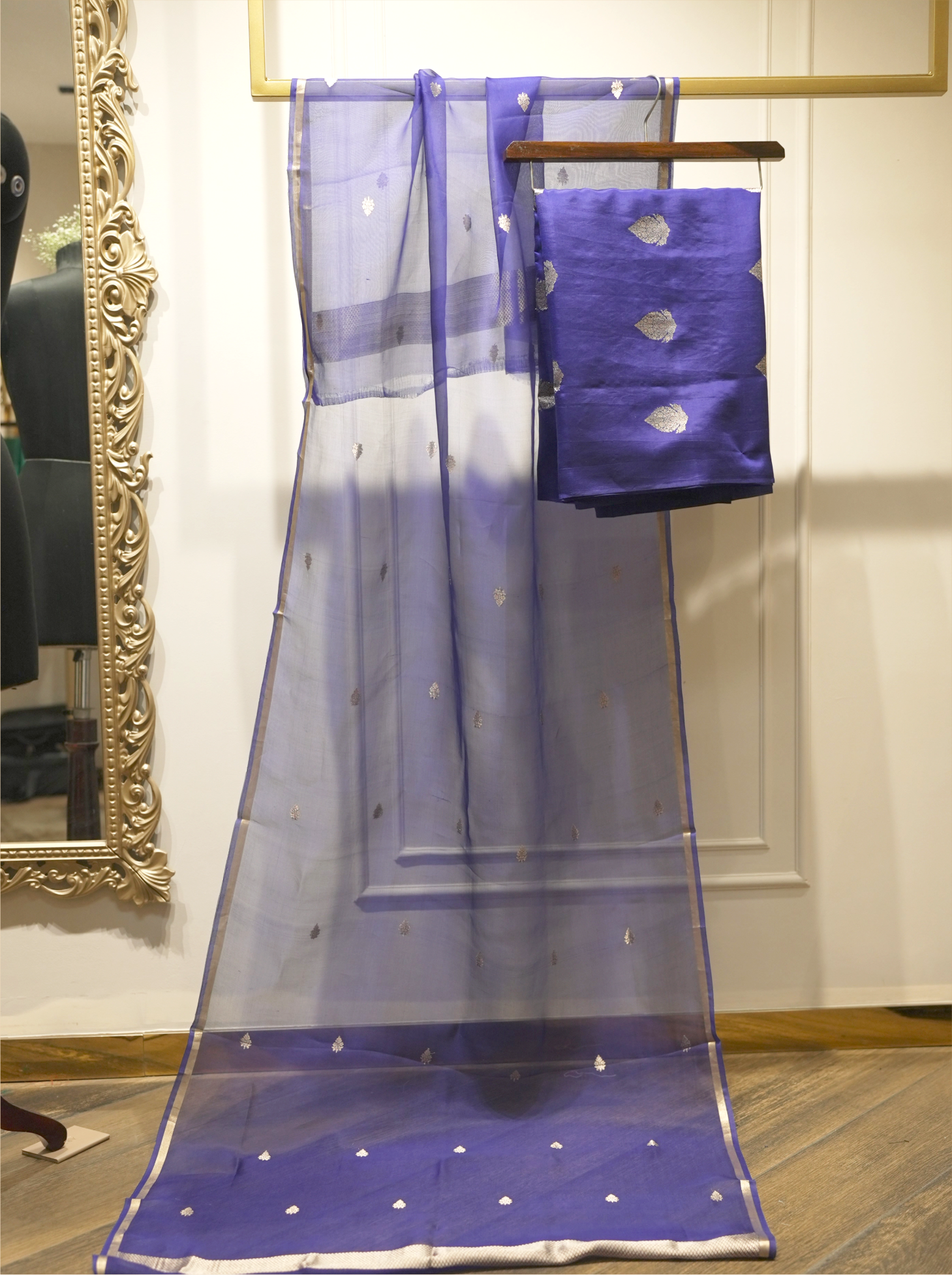 Sheer royal blue handwoven silk dupatta with silver zari bootis and tissue borders, displayed vertically alongside folded suit fabric on a wooden hanger near an ornate gold mirror