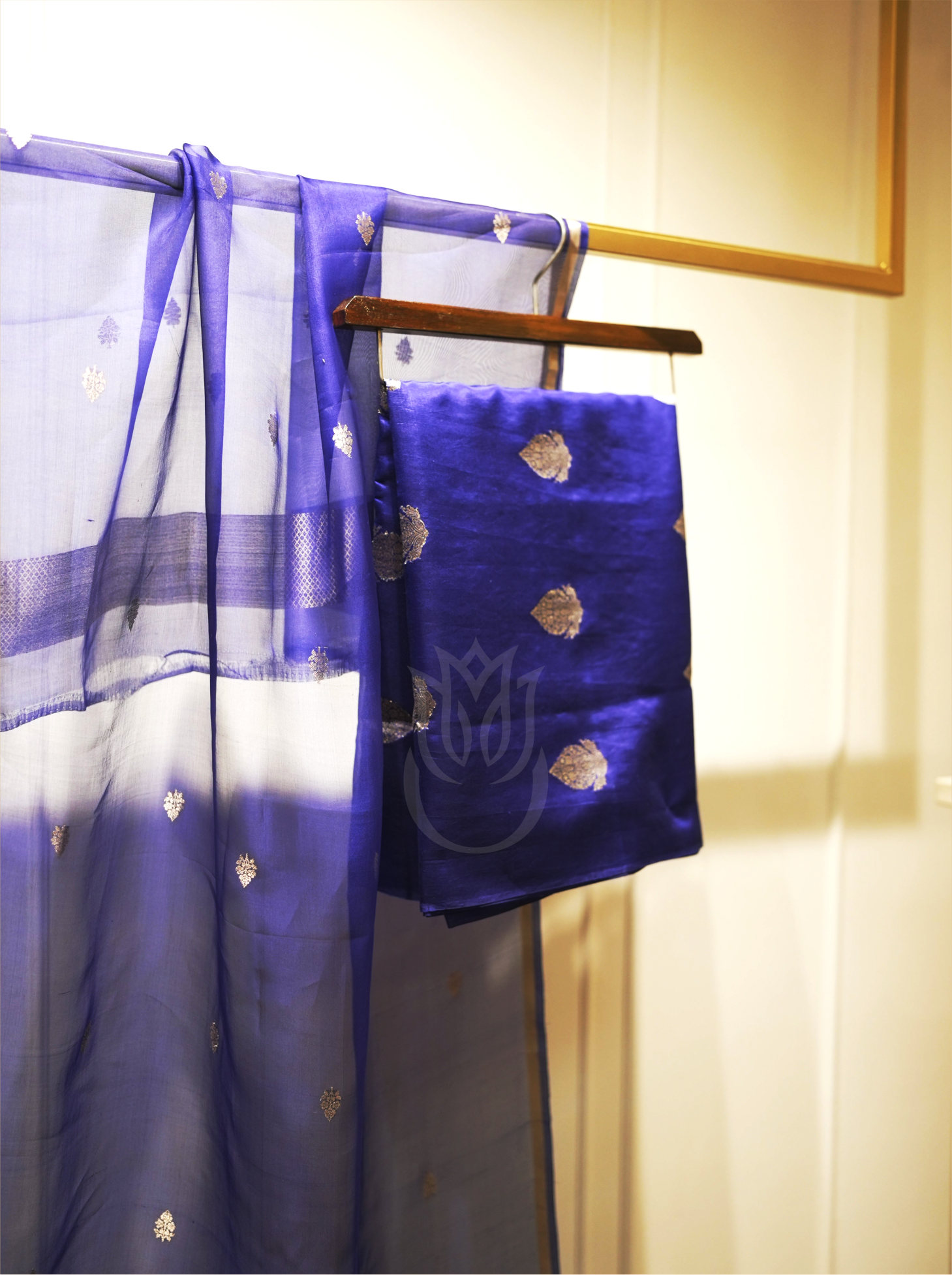 Royal blue handwoven silk suit fabric with gold zari motifs folded on a wooden hanger, paired with a matching sheer dupatta featuring silver zari bootis and tissue borders, set near an ornate gold mirror