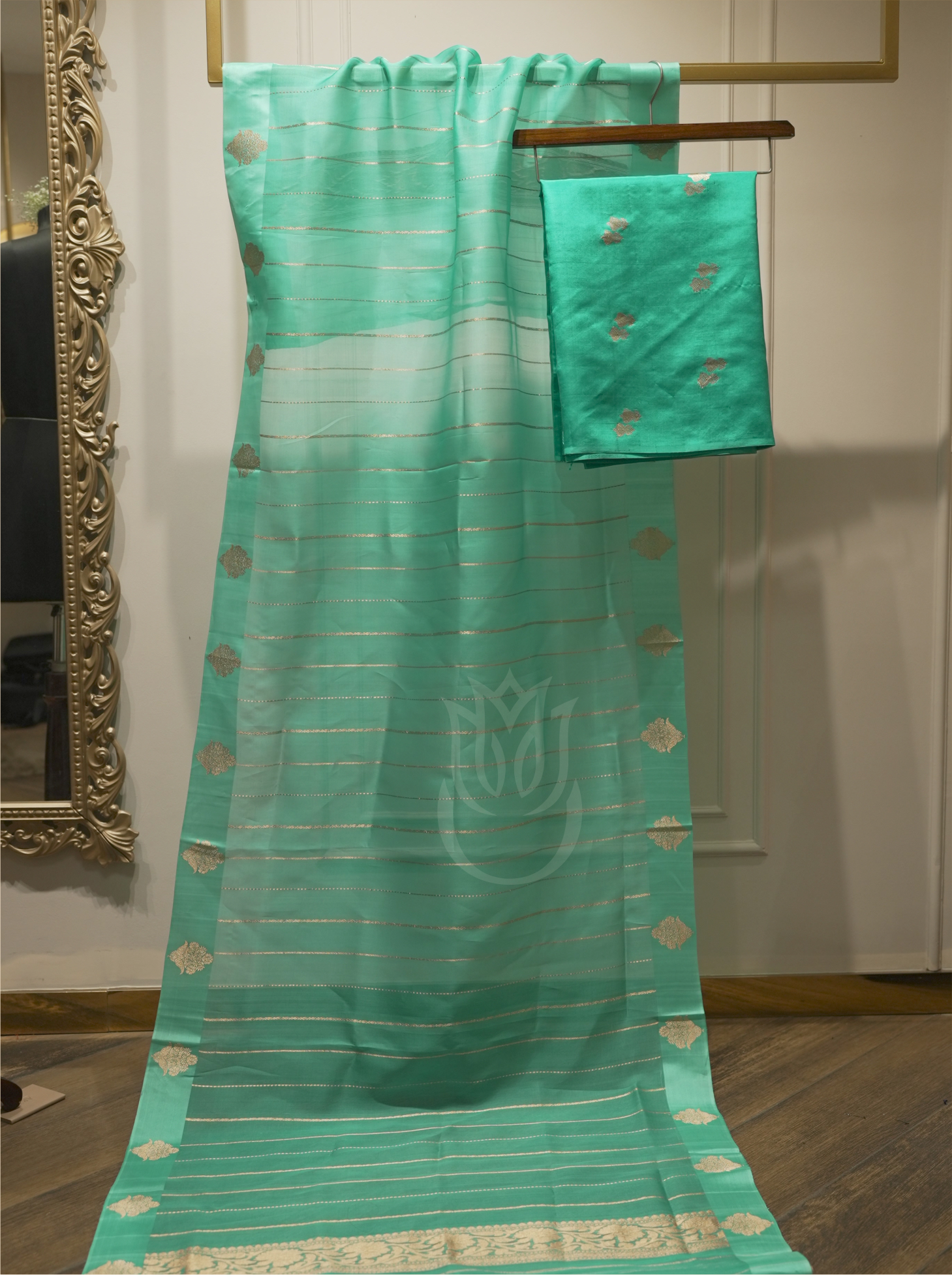 Sheer seafoam green handwoven silk dupatta with gold zari striped pattern and geometric motifs, displayed vertically alongside folded suit fabric on a wooden hanger near an ornate gold mirror