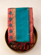 Neatly folded peacock teal green handwoven Paithani silk saree placed inside a traditional brass urli bowl alongside a stack of matching green and red glass bangles, showcasing its crimson red zari woven border with floral and geometric motifs.
