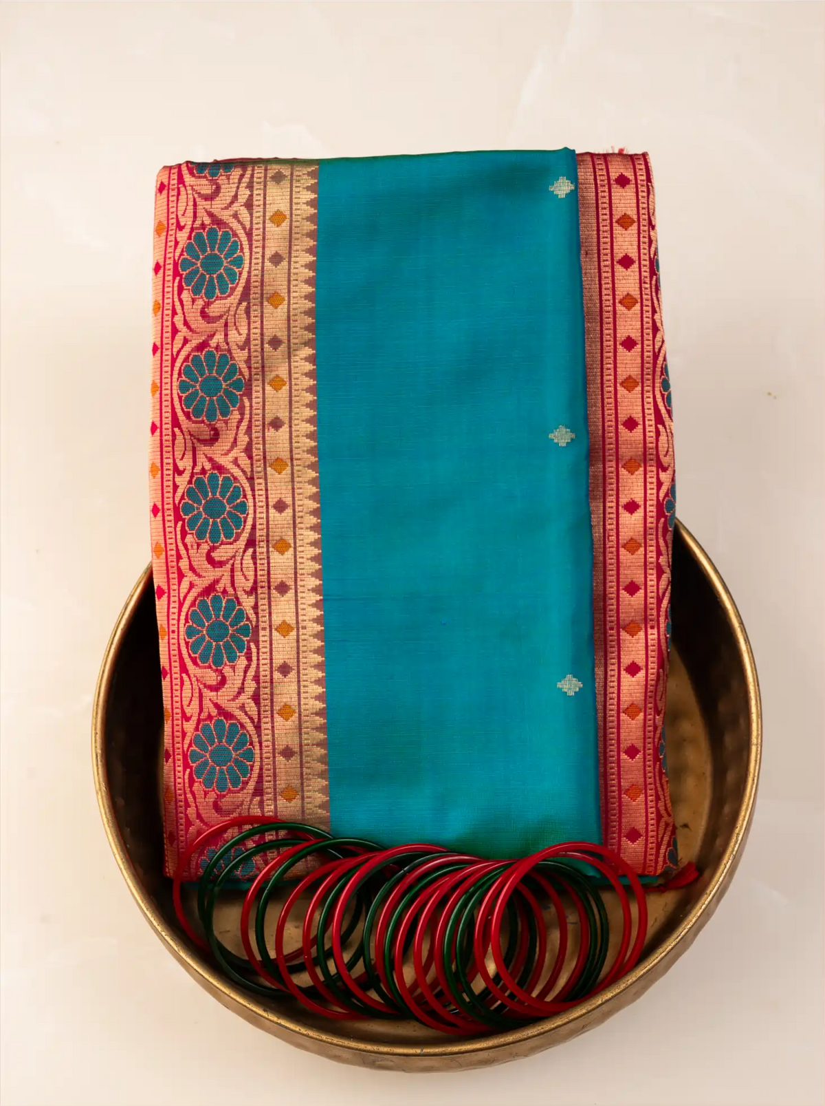 Neatly folded peacock teal green handwoven Paithani silk saree placed inside a traditional brass urli bowl alongside a stack of matching green and red glass bangles, showcasing its crimson red zari woven border with floral and geometric motifs.