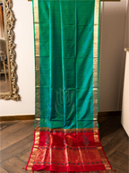 Full-length view of a handwoven peacock teal green Paithani silk saree hung vertically against a wooden floor background, featuring small gold bootis across the main body, slender gold zari borders along the sides, and a broad crimson red pallu at the bottom adorned with intricate gold zari paisley and floral motifs.