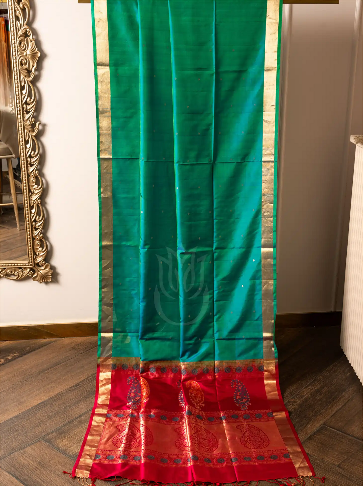 Full-length view of a handwoven peacock teal green Paithani silk saree hung vertically against a wooden floor background, featuring small gold bootis across the main body, slender gold zari borders along the sides, and a broad crimson red pallu at the bottom adorned with intricate gold zari paisley and floral motifs.
