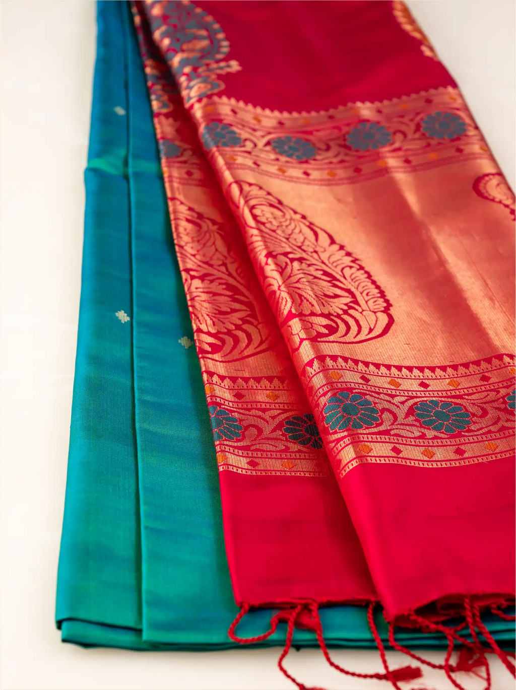 A neatly folded handwoven teal green Paithani silk saree showcasing small gold motifs along the body, juxtaposed against a broad, rich crimson red pallu with gold zari weaving, detailed geometric border bands, and fine red tassels at the edge.
