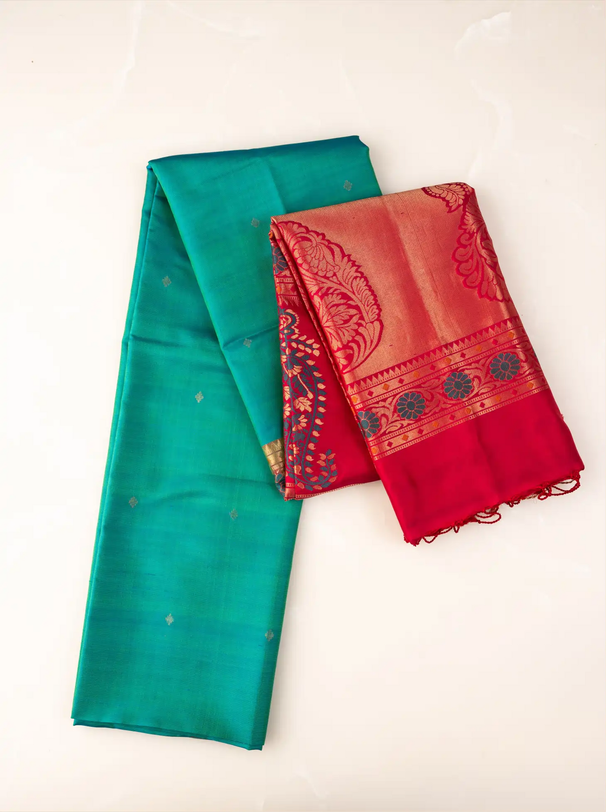 Neatly folded handwoven peacock teal green Paithani silk saree displaying small gold bootis on the body, paired with a folded contrast red zari pallu featuring woven floral bands and peacock motifs with small red fringe tassels.