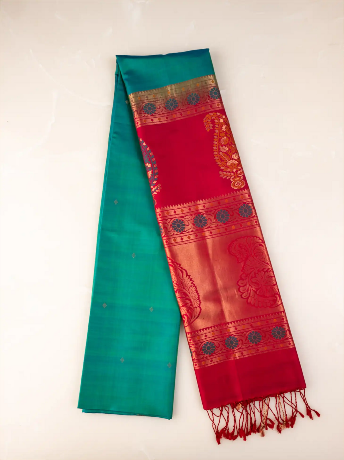Neatly folded handwoven peacock teal green Paithani silk saree showcasing a rich, contrasting crimson red pallu and border adorned with intricate gold zari woven floral and peacock motifs, finished with delicate red tassels at the edge against a light background.