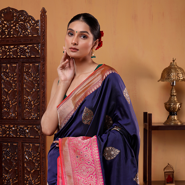 A woman posing in an indigo blue pure Katan silk saree featuring a contrasting pink and gold Sona Rupa boota pallu, standing next to a carved wooden screen.