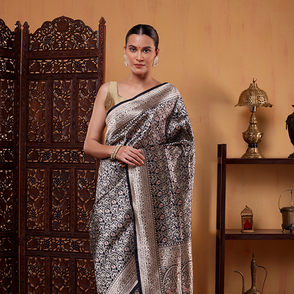 A woman modeling an indigo and gold Banarasi Katan silk saree with an intricate floral Shikargah pattern, styled with a gold blouse.