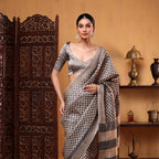 A woman posing in a natural ivory pure Tussar silk saree with delicate all-over boota embroidery work and a black-and-grey striped border next to a carved wooden screen.