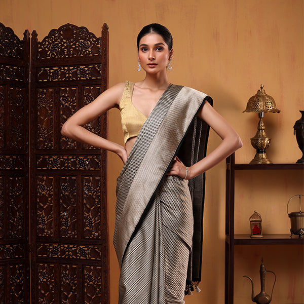 Three-quarter view of a woman modeling the Ivory Noir Tanchoi Banarasi Silk Saree with both hands on her waist, highlighting the pleated drape and contrast gold blouse.