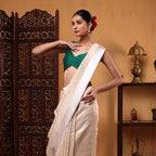 A woman posing in an ivory-colored handwoven Banarasi Munga silk saree featuring a delicate white satin border and a contrasting green sleeveless blouse.