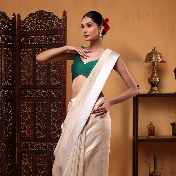 A woman posing in an ivory-colored handwoven Banarasi Munga silk saree featuring a delicate white satin border and a contrasting green sleeveless blouse.
