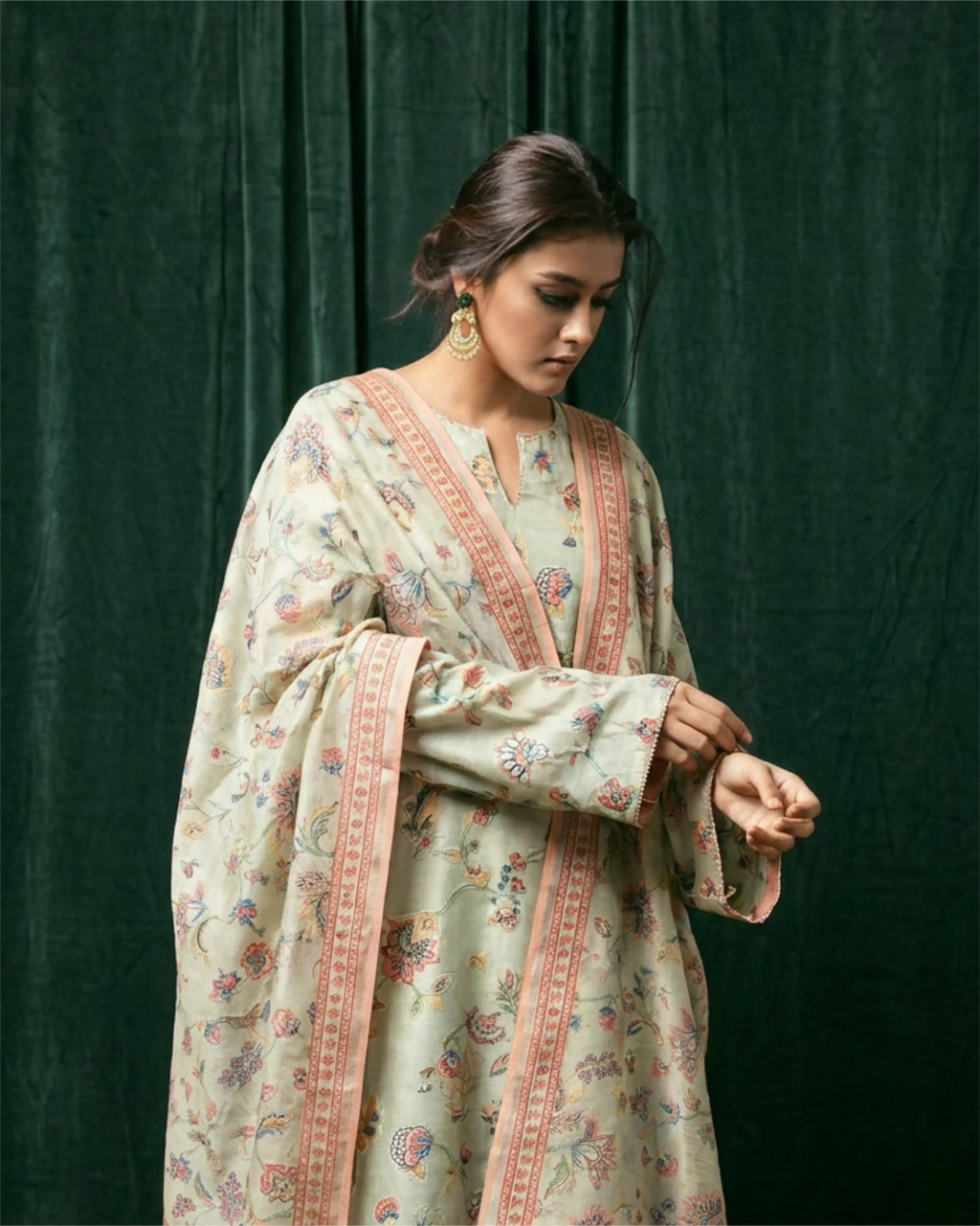 A woman wearing a light beige floral-printed kurta with delicate pink border trim, standing against a sophisticated dark green velvet curtain backdrop.