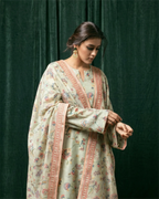 A woman wearing a light beige floral-printed kurta with delicate pink border trim, standing against a sophisticated dark green velvet curtain backdrop.