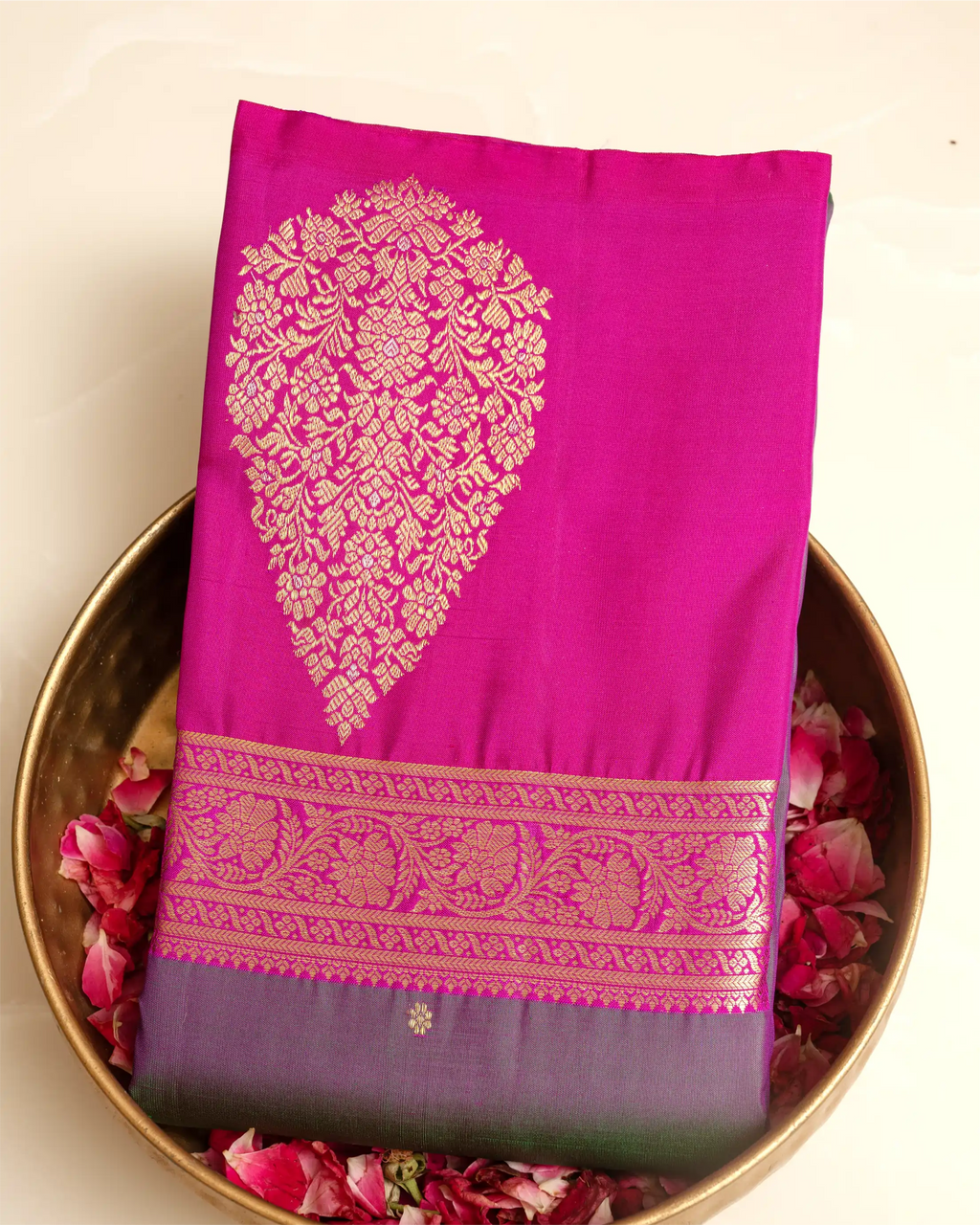 A folded magenta pink silk saree with a large gold floral zari motif and a decorative gold border, displayed on a gold-colored plate filled with pink rose petals