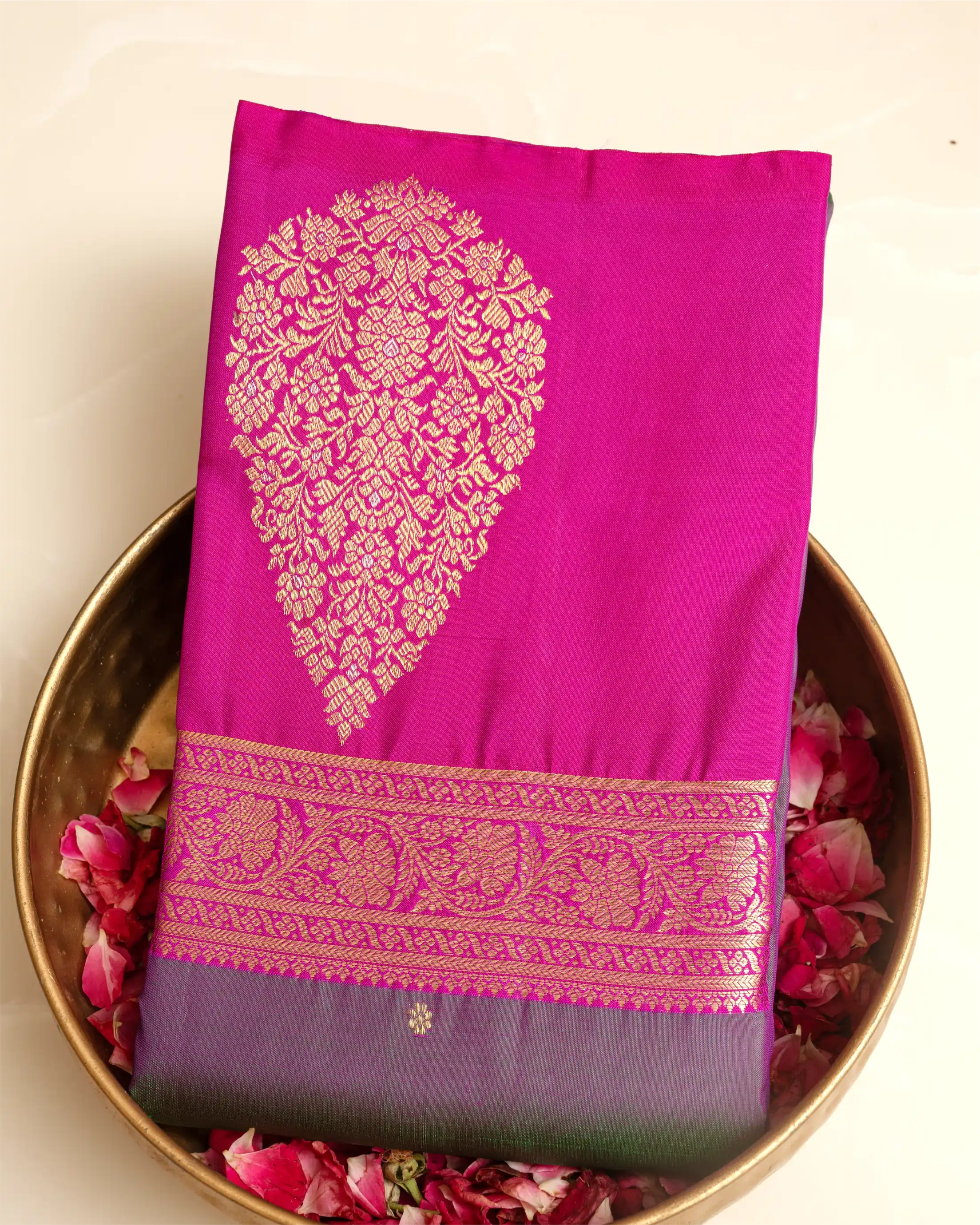 A folded magenta pink silk saree with a large gold floral zari motif and a decorative gold border, displayed on a gold-colored plate filled with pink rose petals