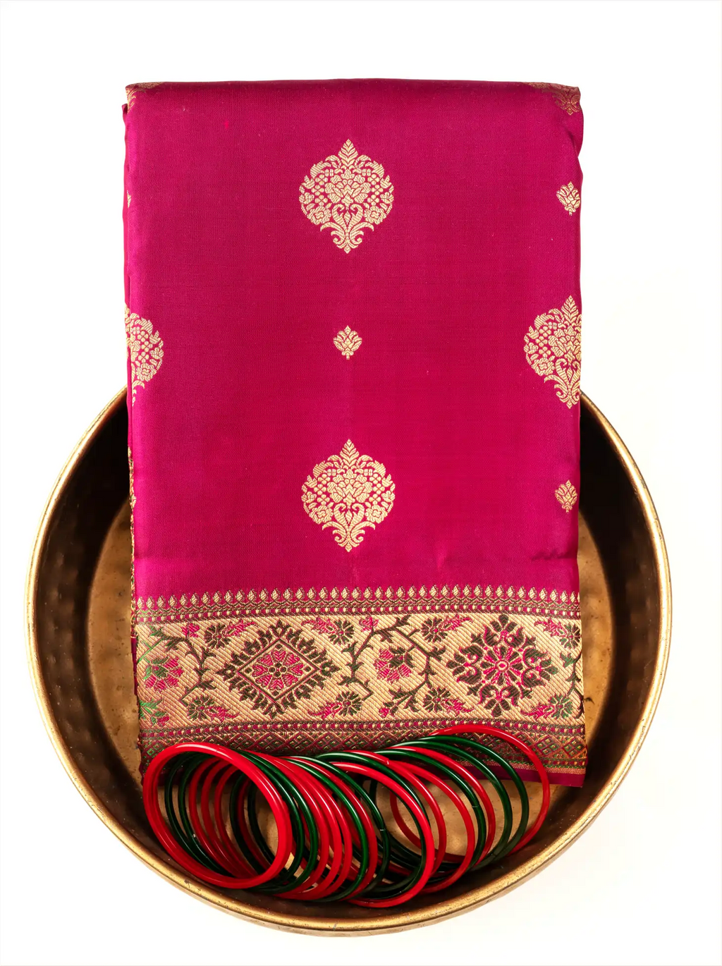 A neatly folded bright magenta-pink Kanjivaram Paithani silk saree arranged inside a traditional round brass plate, featuring woven gold zari floral medallions on the fabric, a decorated gold zari border with pink motif accents, and set with red and green glass bangles in front.