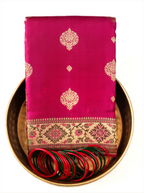 A neatly folded bright magenta-pink Kanjivaram Paithani silk saree arranged inside a traditional round brass plate, featuring woven gold zari floral medallions on the fabric, a decorated gold zari border with pink motif accents, and set with red and green glass bangles in front.