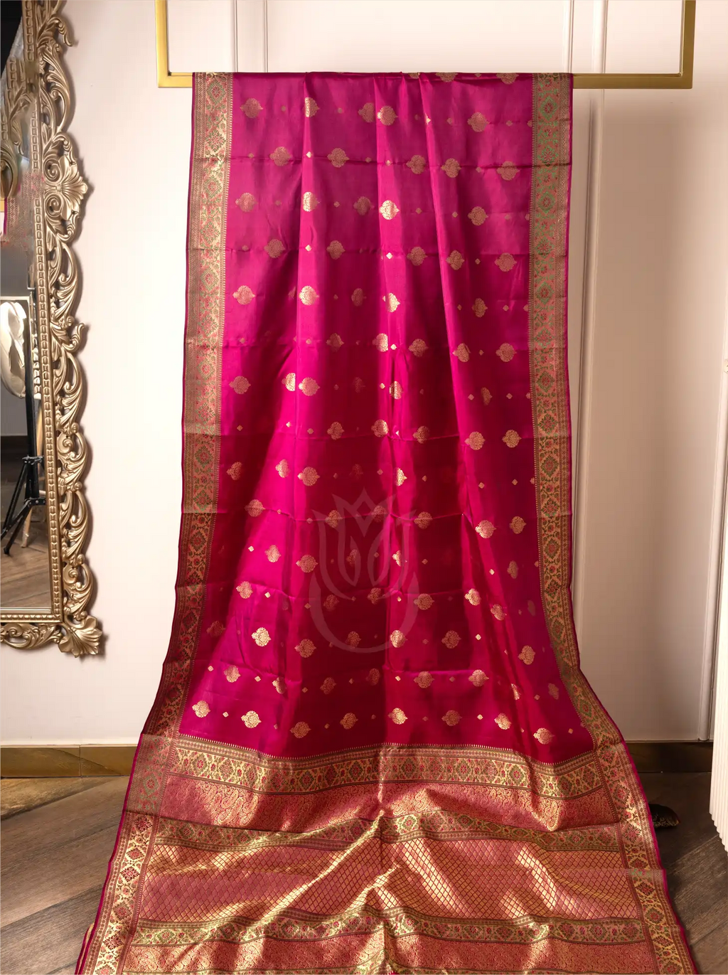 A vibrant magenta-pink handwoven pure silk Kanjivaram Paithani saree displayed on a hanging frame, featuring small woven gold zari floral butas across the body, a central lotus motif watermarked on the fabric, and a rich, wide gold zari woven border and pallu.
