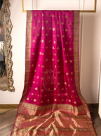 A vibrant magenta-pink handwoven pure silk Kanjivaram Paithani saree displayed on a hanging frame, featuring small woven gold zari floral butas across the body, a central lotus motif watermarked on the fabric, and a rich, wide gold zari woven border and pallu.