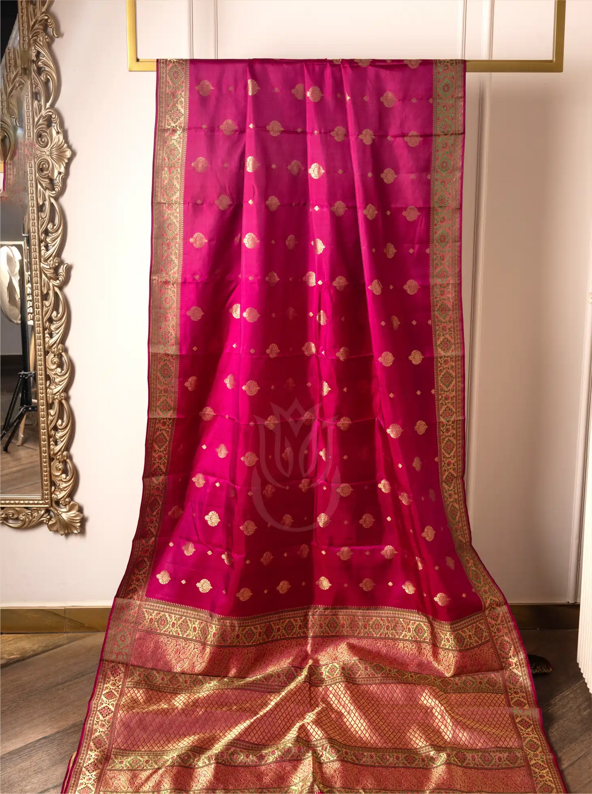 A vibrant magenta-pink handwoven pure silk Kanjivaram Paithani saree displayed on a hanging frame, featuring small woven gold zari floral butas across the body, a central lotus motif watermarked on the fabric, and a rich, wide gold zari woven border and pallu.