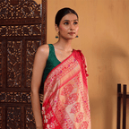 A woman wearing the Majestic Magenta pure Katan silk saree featuring a handwoven golden Zari Paithani border, styled with a contrasting dark green sleeveless blouse near a wooden screen.