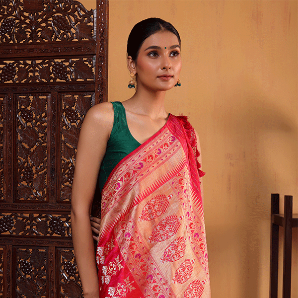 A woman wearing the Majestic Magenta pure Katan silk saree featuring a handwoven golden Zari Paithani border, styled with a contrasting dark green sleeveless blouse near a wooden screen.