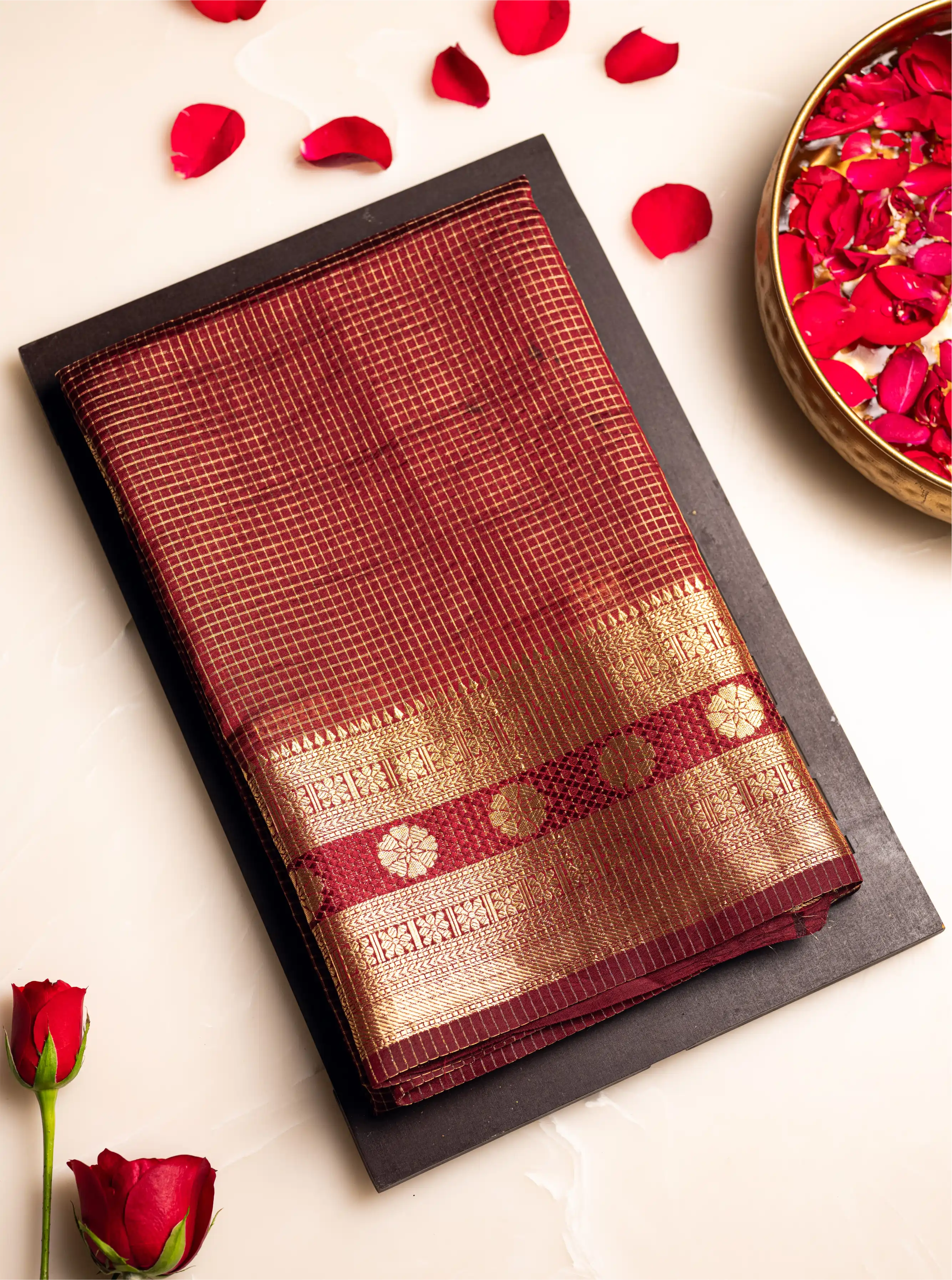 Flat-lay view of a folded maroon Handwoven Pure Katan Silk Saree featuring a gold zari border and small floral butas, decorated with red rose petals.