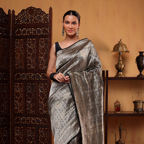 A woman modeling a black and silver Banarasi silk saree with opulent golden zari stripes, standing by a traditional wooden screen.