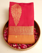 A folded orange and pink Kanjivaram silk saree with an intricate gold zari brocade motif and border, displayed in a brass bowl filled with fresh pink rose petals.