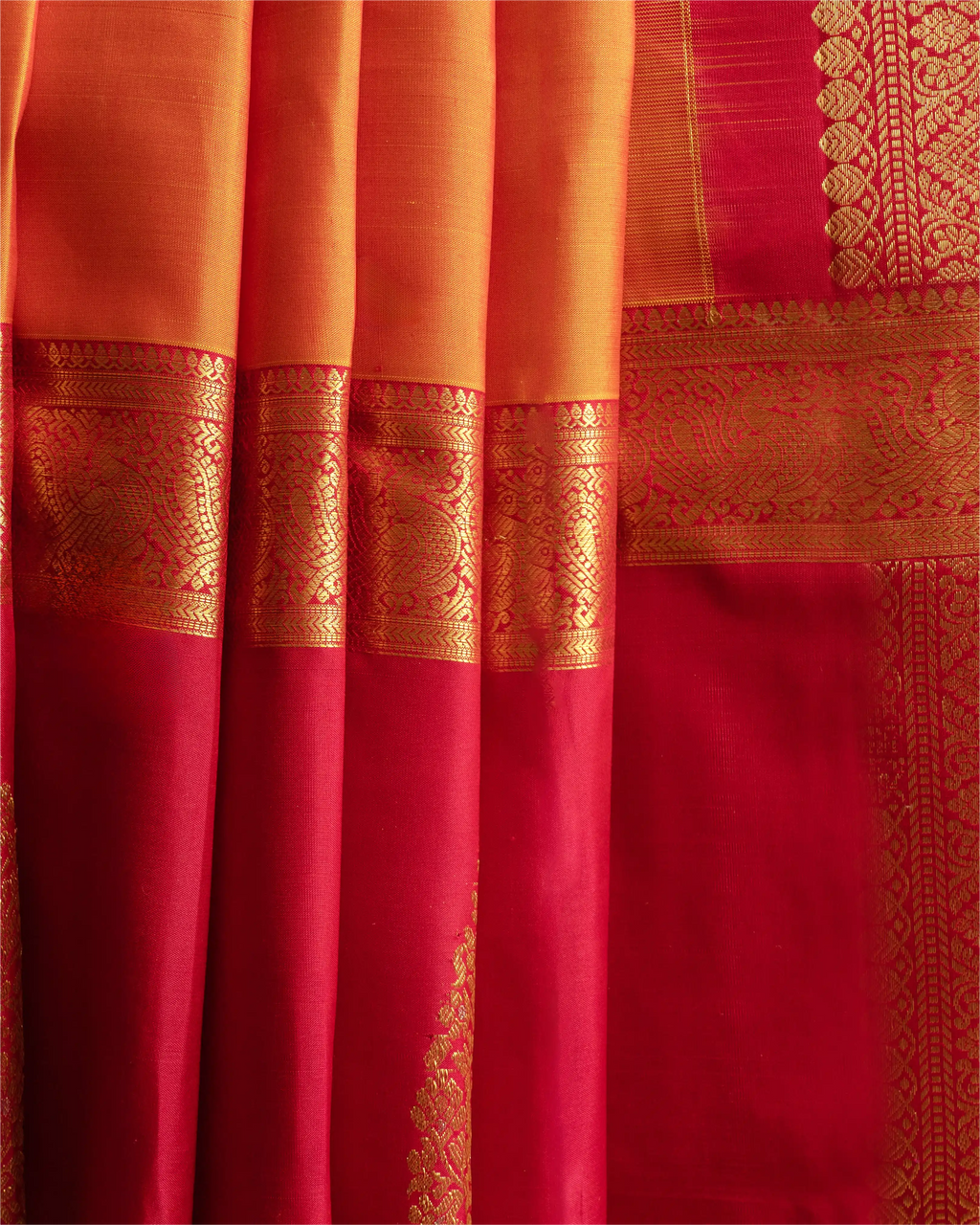Close-up of neatly pleated orange and red Kanjivaram silk saree fabric highlighting the intricate gold zari woven borders and rich texture