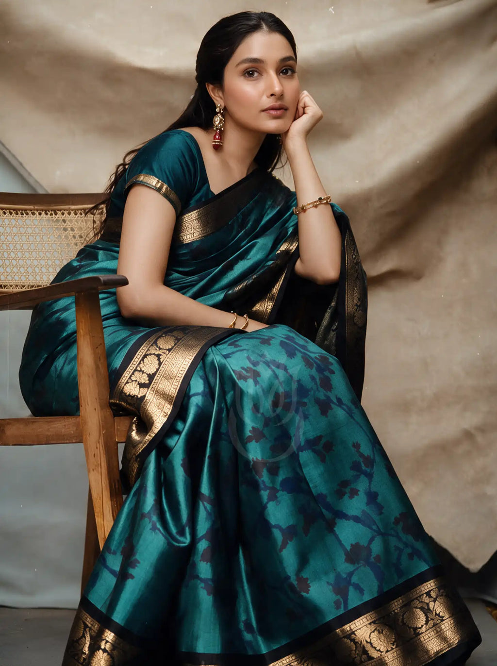 Model wearing a peacock blue and teal handwoven Patola Jacquard silk saree with a dark teal blouse, featured sitting on a wooden chair. The saree showcases intricate woven jacquard motifs with a broad gold zari border along the pallu.