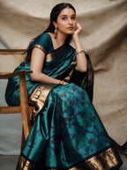 Model wearing a peacock blue and teal handwoven Patola Jacquard silk saree with a dark teal blouse, featured sitting on a wooden chair. The saree showcases intricate woven jacquard motifs with a broad gold zari border along the pallu.