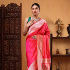 A woman modeling a pink pure handwoven Banarasi Katan silk saree with an intricate gold Shikargah pallu and a green blouse, standing in front of a traditional wooden screen.
