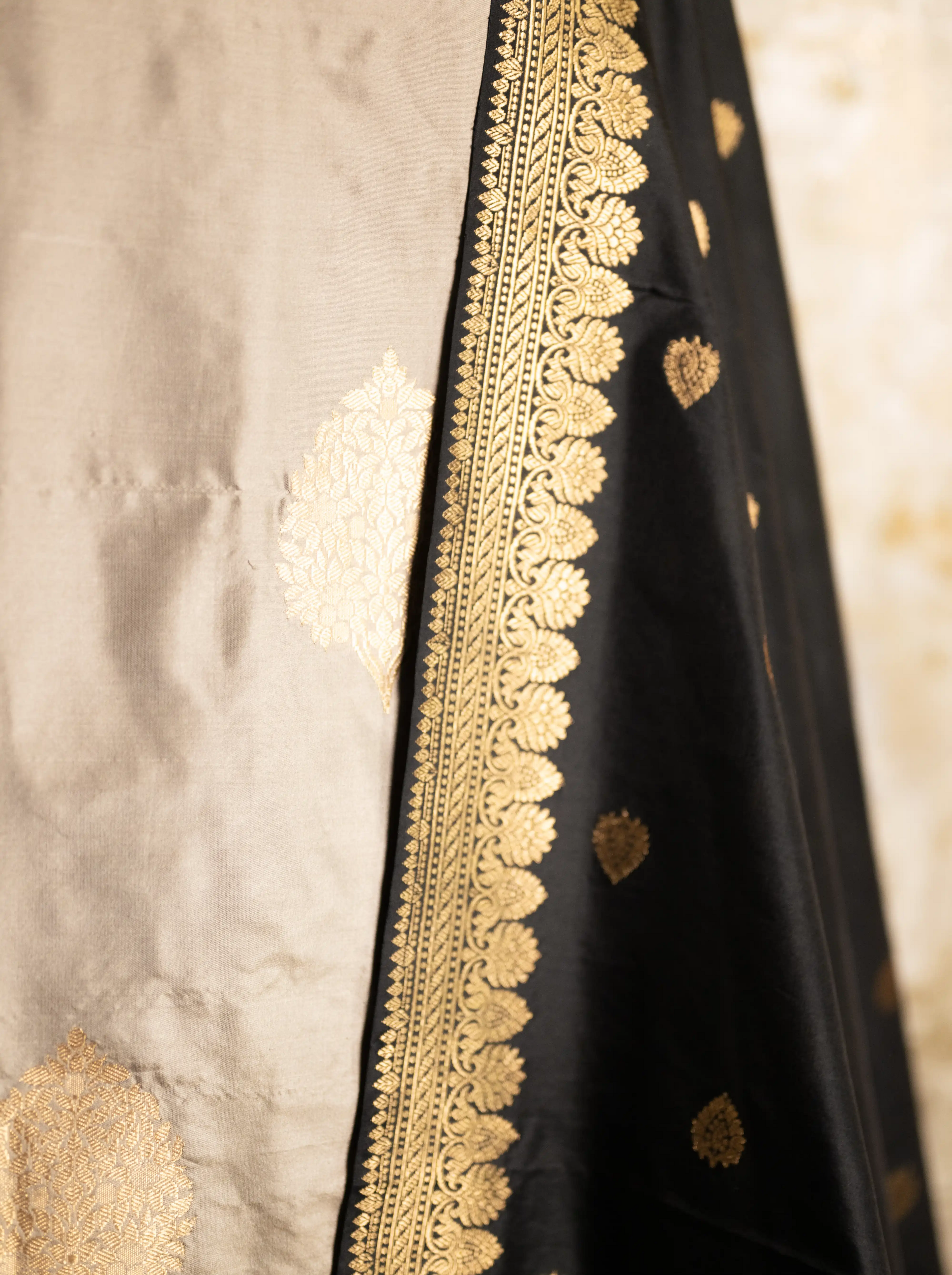 Close-up of intricate gold zari embroidery on a black Katan silk dupatta with a textured beige background.